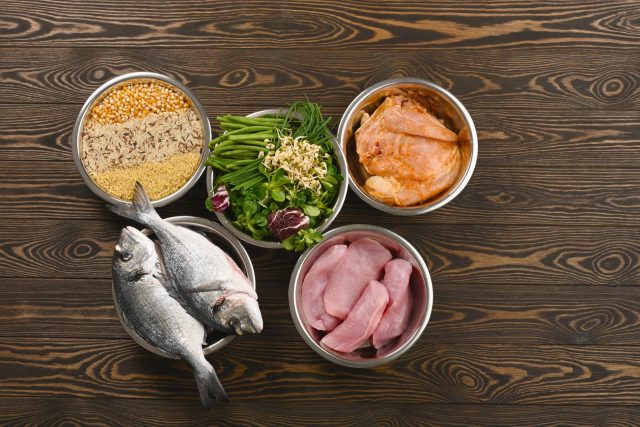 Bowls of raw dog food ingredients including chicken breast, whole fish, grains, and fresh greens on a wood table
